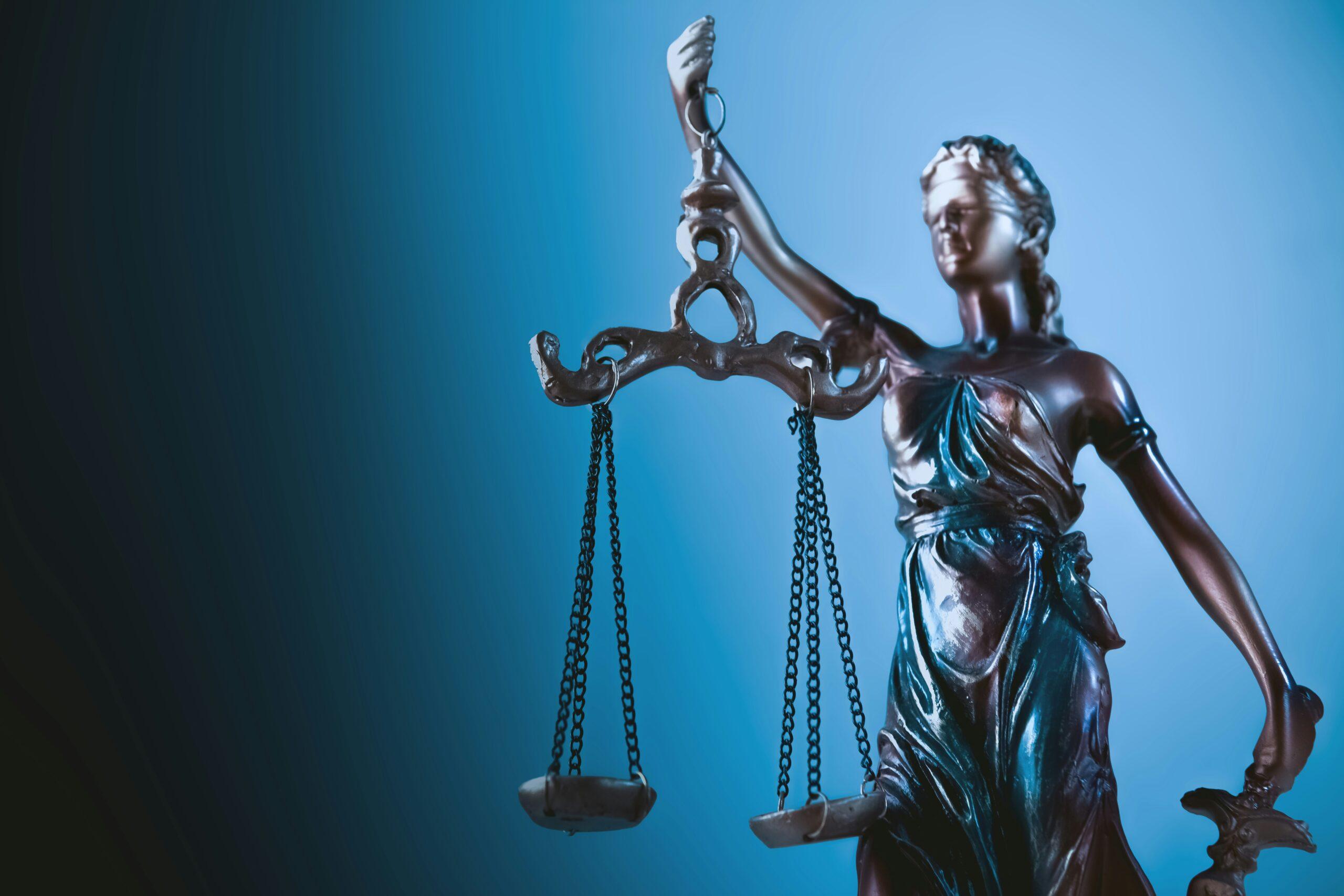Bronze statue of Lady Justice holding a set of balanced scales in her raised hand, illuminated against a blue gradient background, symbolizing law, fairness, and judicial authority, with detailed drapery and metal chains visible on the scales.