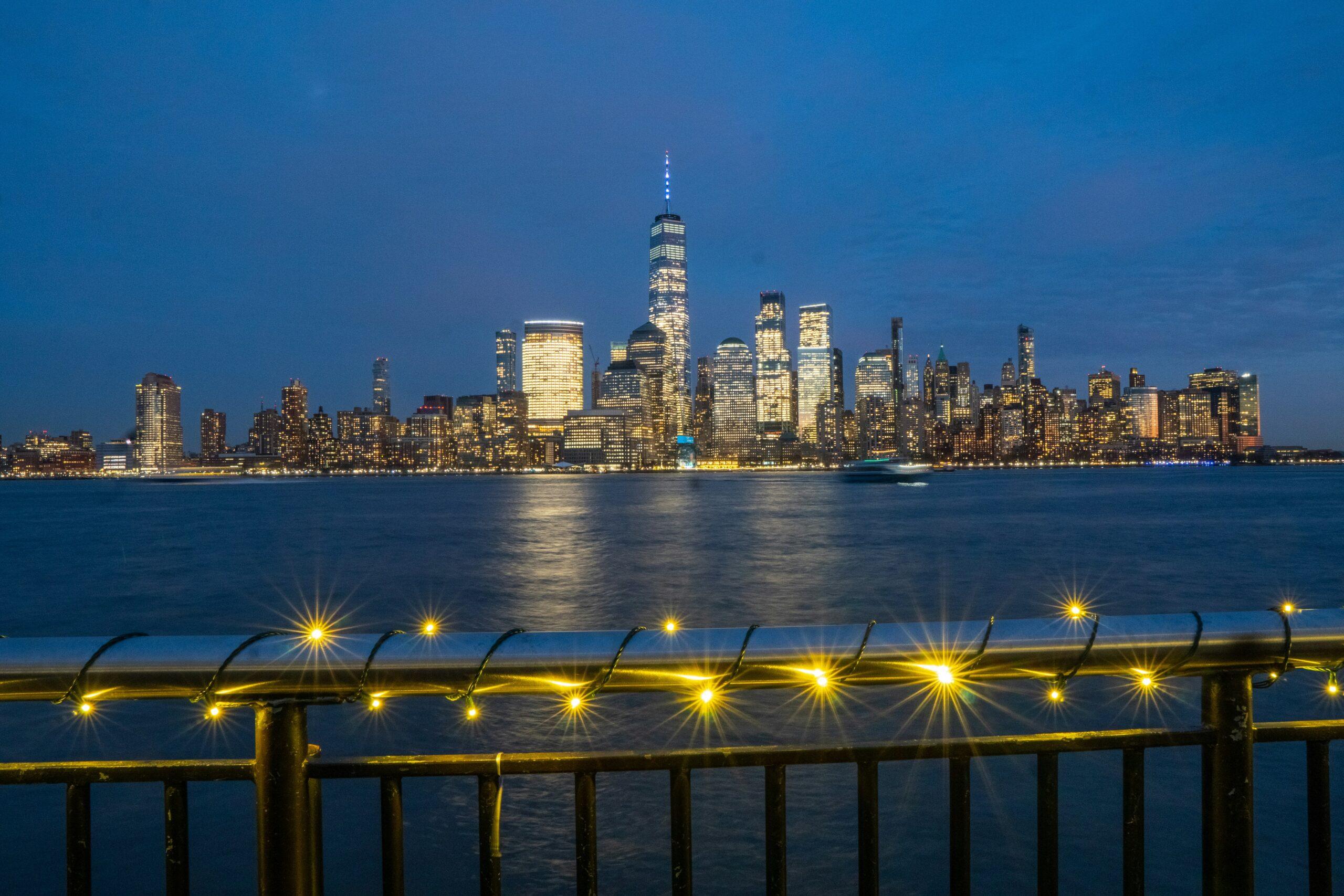Nighttime city skyline viewed across a river, with illuminated skyscrapers reflecting on the water and a waterfront railing in the foreground decorated with glowing string lights.