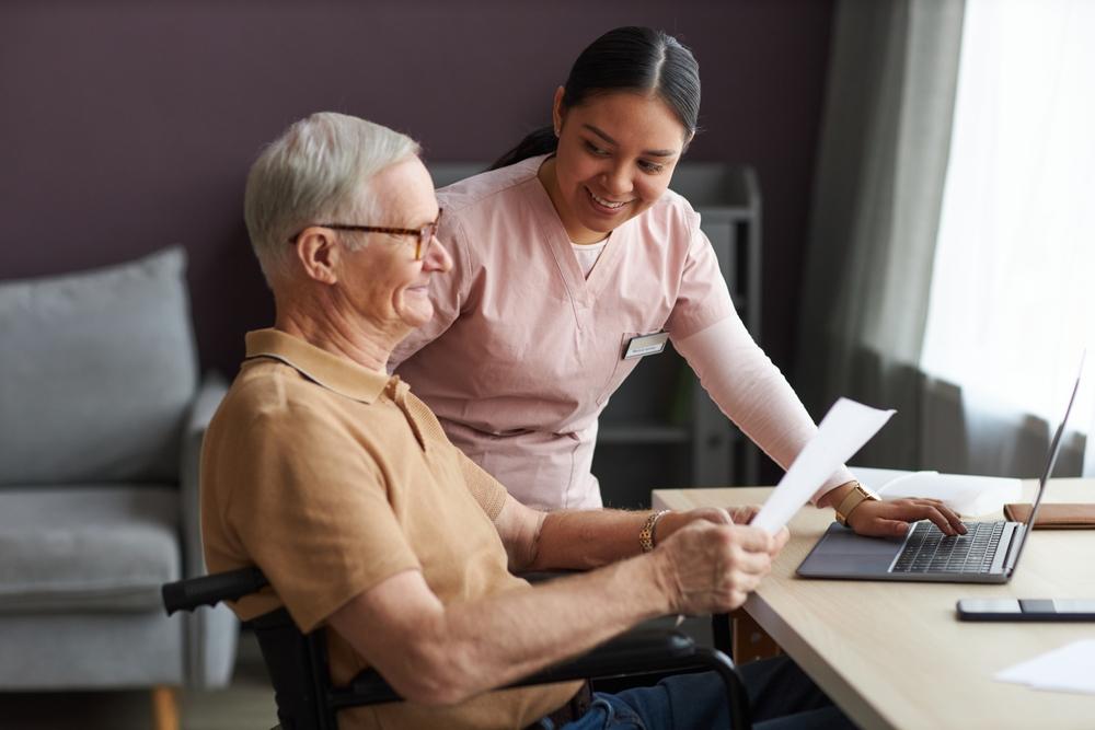 Caregiver assisting an elderly man while reviewing paperwork or documents together at home or in a care facility setting